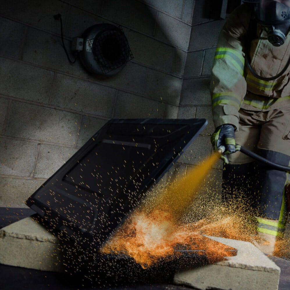 A laptop on fire with bright flames and smoke, while a person uses a fire extinguisher to put out the blaze.